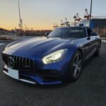 Blue Mercedes convertible in a parking lot at sunset, with cranes and a shipyard in the background
