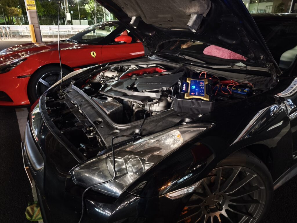 Car with hood open showing a complex engine bay and a portable jump starter connected nearby in a dimly lit parking area with a red Ferrari parked beside it? (Describe the setup: engine bay and jump starter).