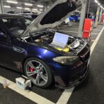 Blue BMW with its hood raised in a parking garage, laptop connected to the engine for diagnostics amidst tools and cables.