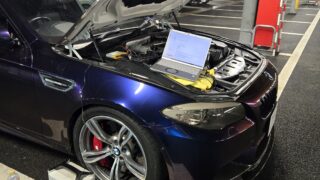 Blue BMW with its hood raised in a parking garage, laptop connected to the engine for diagnostics amidst tools and cables.
