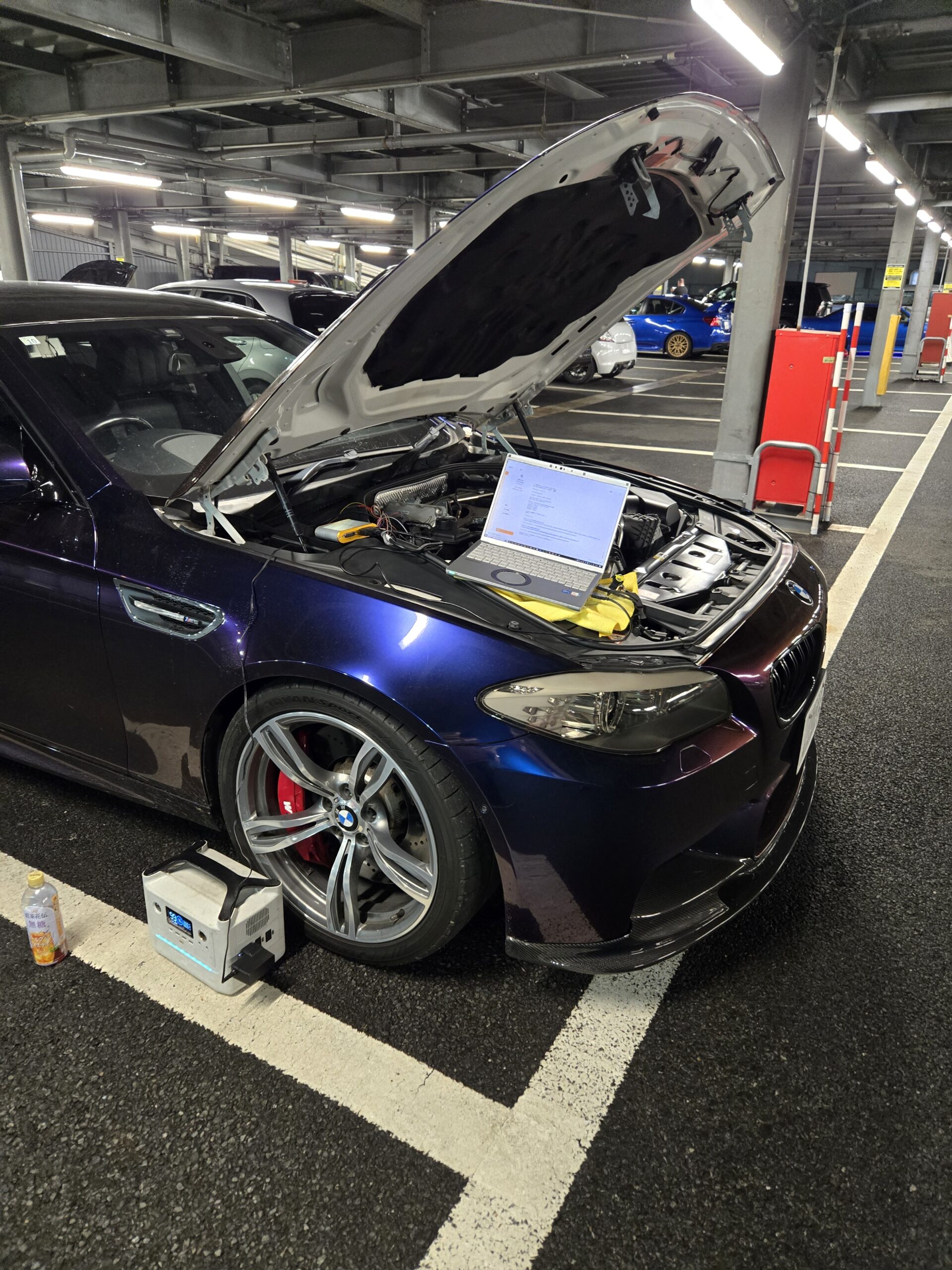 Blue BMW with its hood raised in a parking garage, laptop connected to the engine for diagnostics amidst tools and cables.