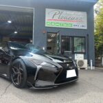 Shiny black Lexus sports car parked in front of a blue auto shop with a 'Pleasure' sign above the entrance