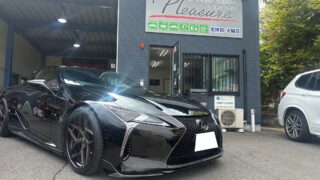 Shiny black Lexus sports car parked in front of a blue auto shop with a 'Pleasure' sign above the entrance