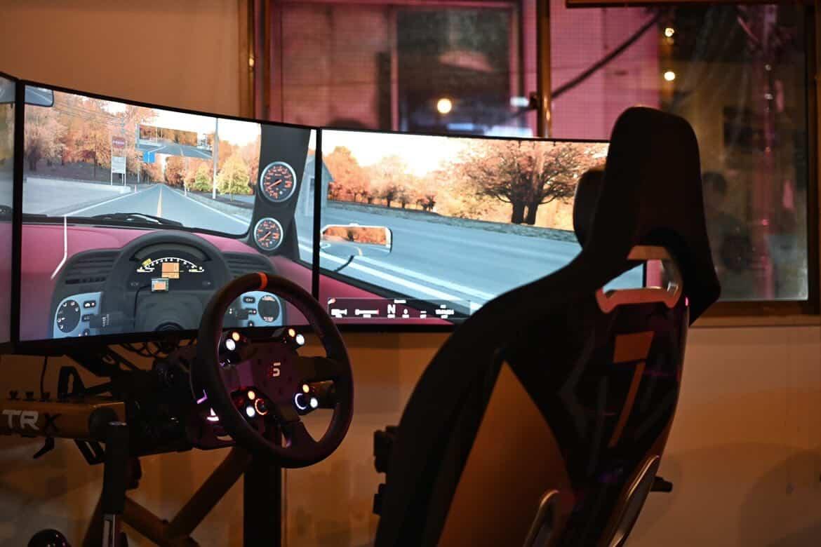 Racing simulator setup with three curved screens showing a road scene, a steering wheel, and a racing seat.