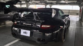 Black sports car with a large rear wing parked in a metal-floored garage.
