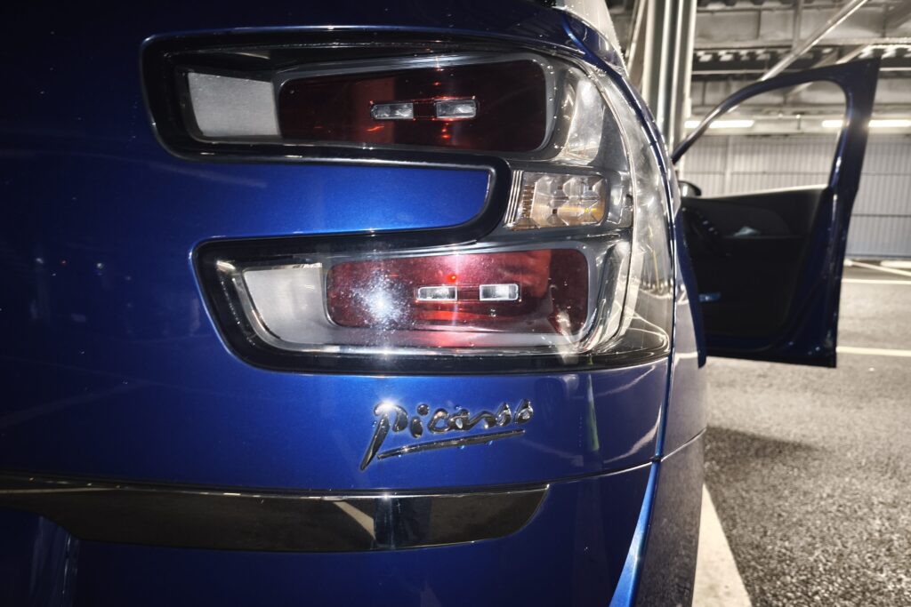 Close-up of a blue car's rear tail lights with the trunk area, a partially open door, and a garage background.