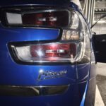 Close-up of a blue car's rear tail lights with the trunk area, a partially open door, and a garage background.