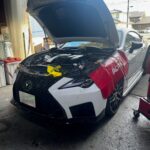 White luxury sedan in a shop with its hood up and engine exposed, tools placed over the front area, cables nearby.