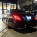 Rear three-quarter view of a black Mercedes-Benz S 500 parked by a storefront at night with two men standing nearby looking at their phones/ chatting.