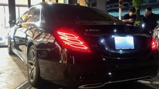 Rear three-quarter view of a black Mercedes-Benz S 500 parked by a storefront at night with two men standing nearby looking at their phones/ chatting.