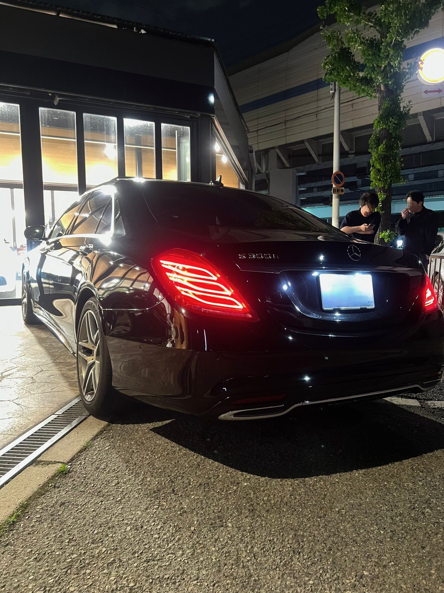 Rear three-quarter view of a black Mercedes-Benz S 500 parked by a storefront at night with two men standing nearby looking at their phones/ chatting.
