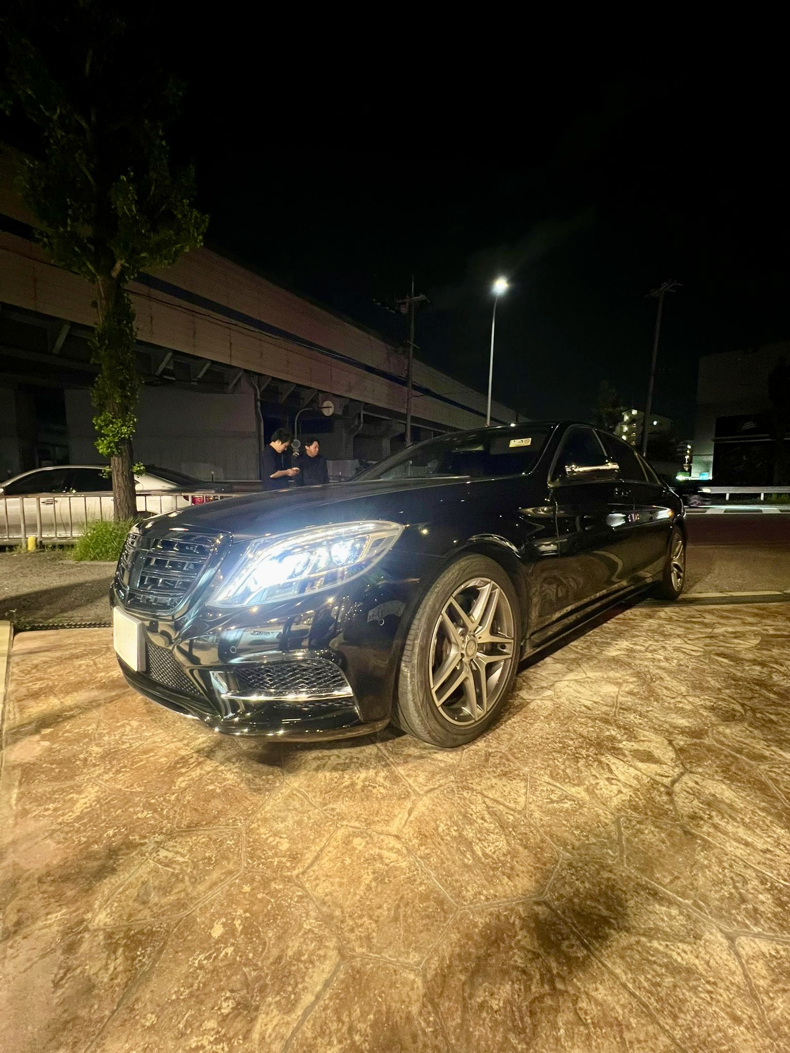Glossy black luxury sedan parked on a lit pavement at night, two men standing nearby looking at a phone or device beside the car.