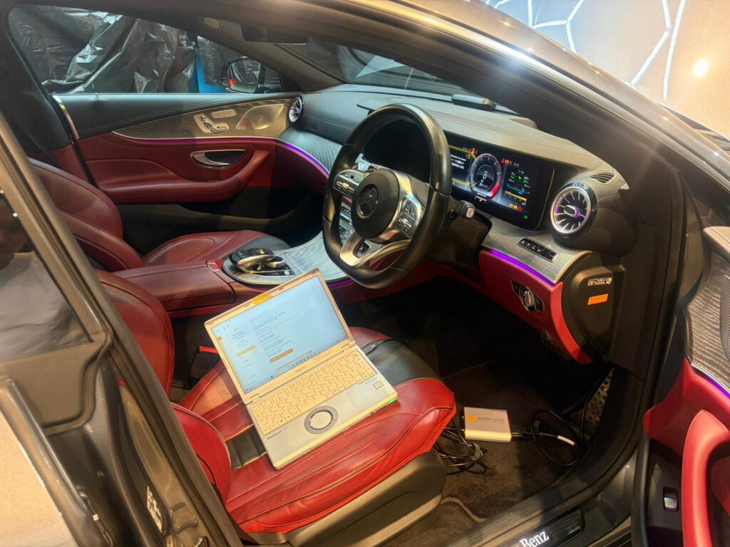 IMG-20260520-WA0010 – mbFAST Tuning Red-leather Mercedes interior with a digital dashboard, purple ambient lighting, and a laptop on the passenger seat edge.