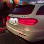 White Mercedes SUV rear view at night with red taillights; colorful shipping containers in the background.