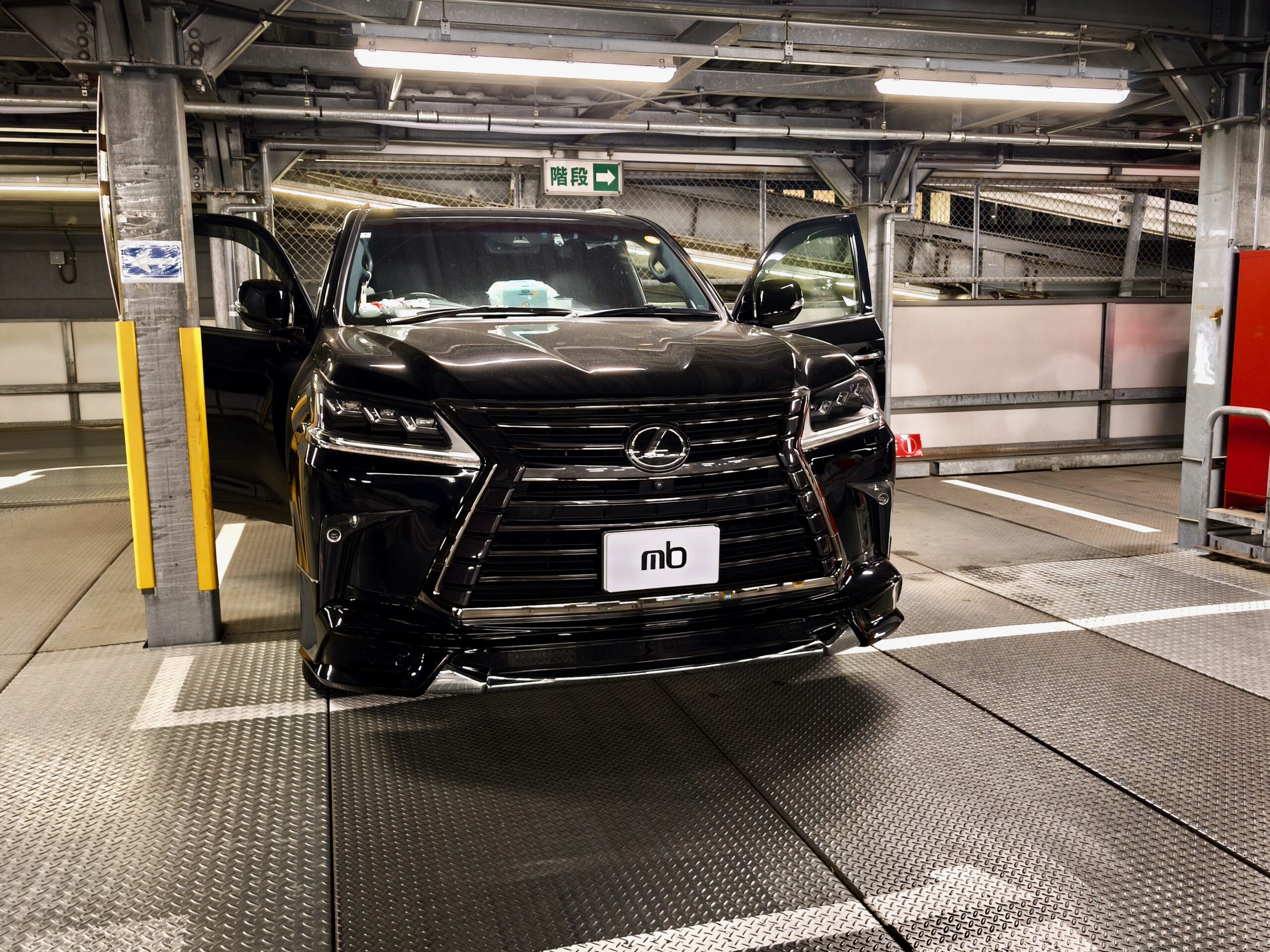 Front view of a shiny black Lexus SUV with both doors open inside an industrial parking garage on a car lift.