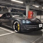 Dark gray Porsche sports car parked in an industrial-looking parking garage with exposed metal beams and bright overhead lights in the background.