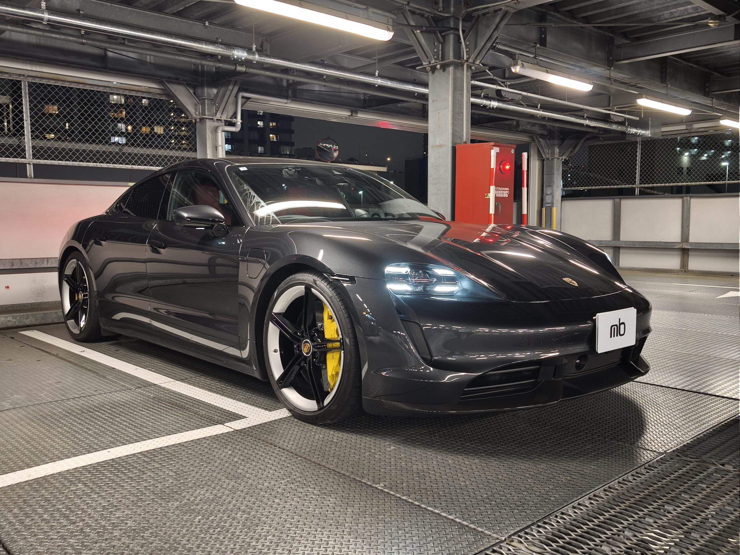 Dark gray Porsche sports car parked in an industrial-looking parking garage with exposed metal beams and bright overhead lights in the background.
