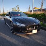Front-left view of a black Maserati on an empty road, with red interior visible and a row of shipping containers in the background.