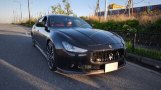Front-left view of a black Maserati on an empty road, with red interior visible and a row of shipping containers in the background.