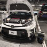Open-engine bay of a white Toyota SUV in a service bay, engine components visible and a diagnostic device on the floor nearby.
