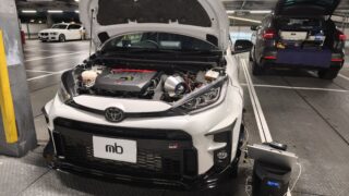 Open-engine bay of a white Toyota SUV in a service bay, engine components visible and a diagnostic device on the floor nearby.