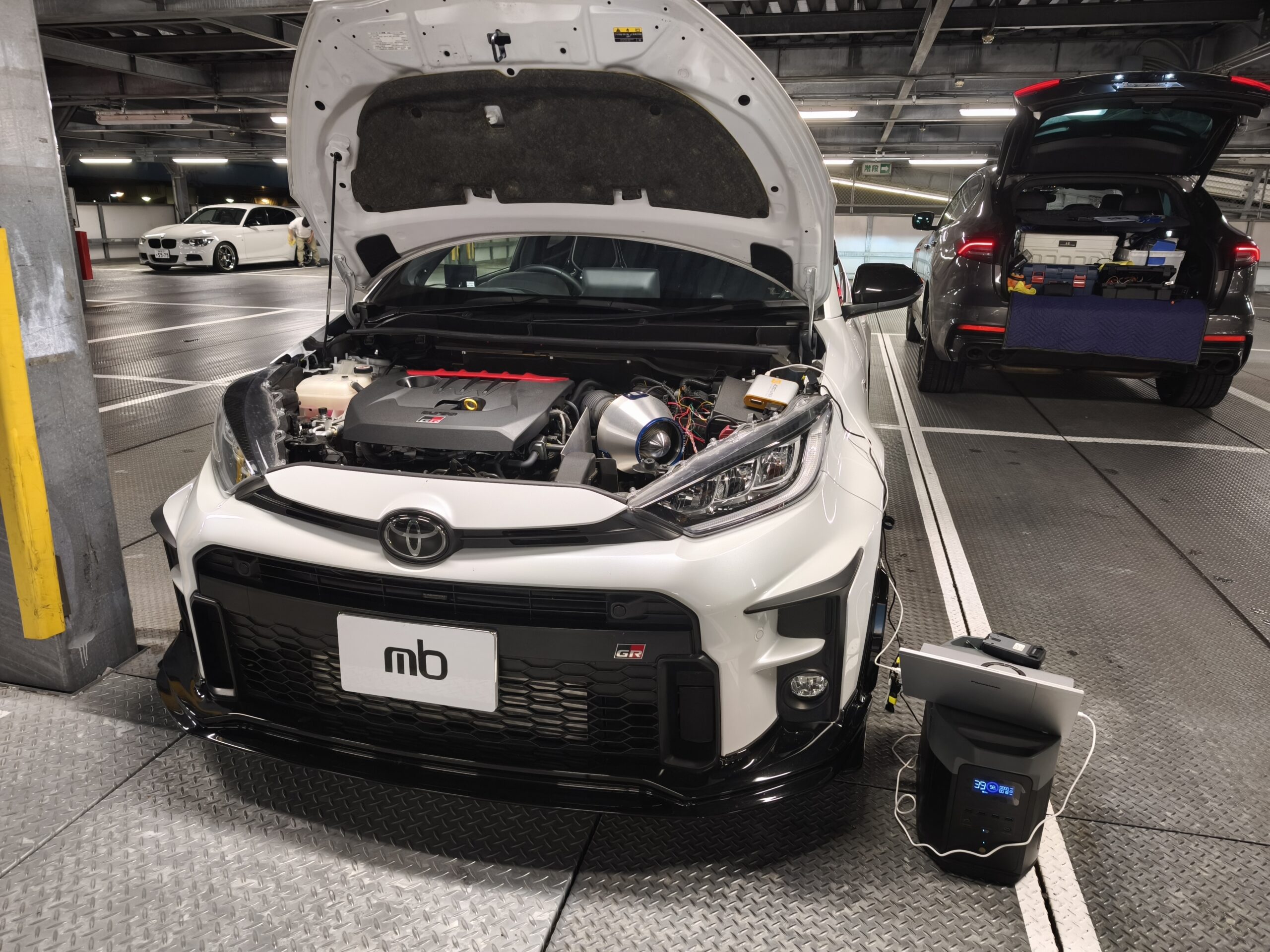 Open-engine bay of a white Toyota SUV in a service bay, engine components visible and a diagnostic device on the floor nearby.