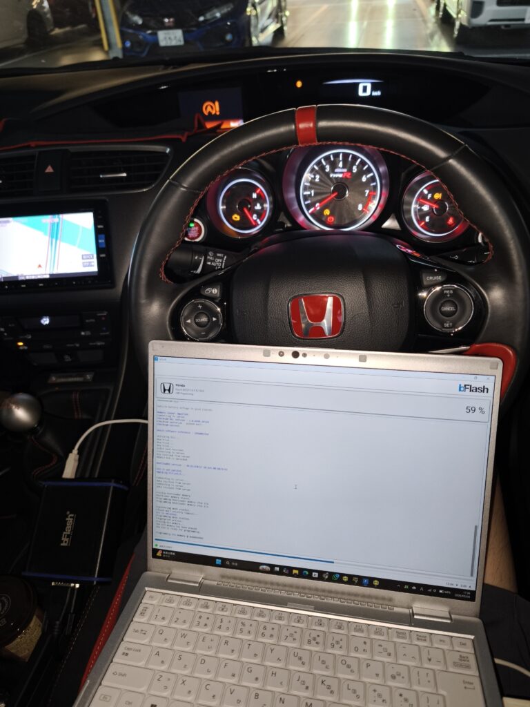 Inside a car cockpit showing the steering wheel and illuminated dashboard gauges with a laptop on the passenger’s lap displaying code or a program.