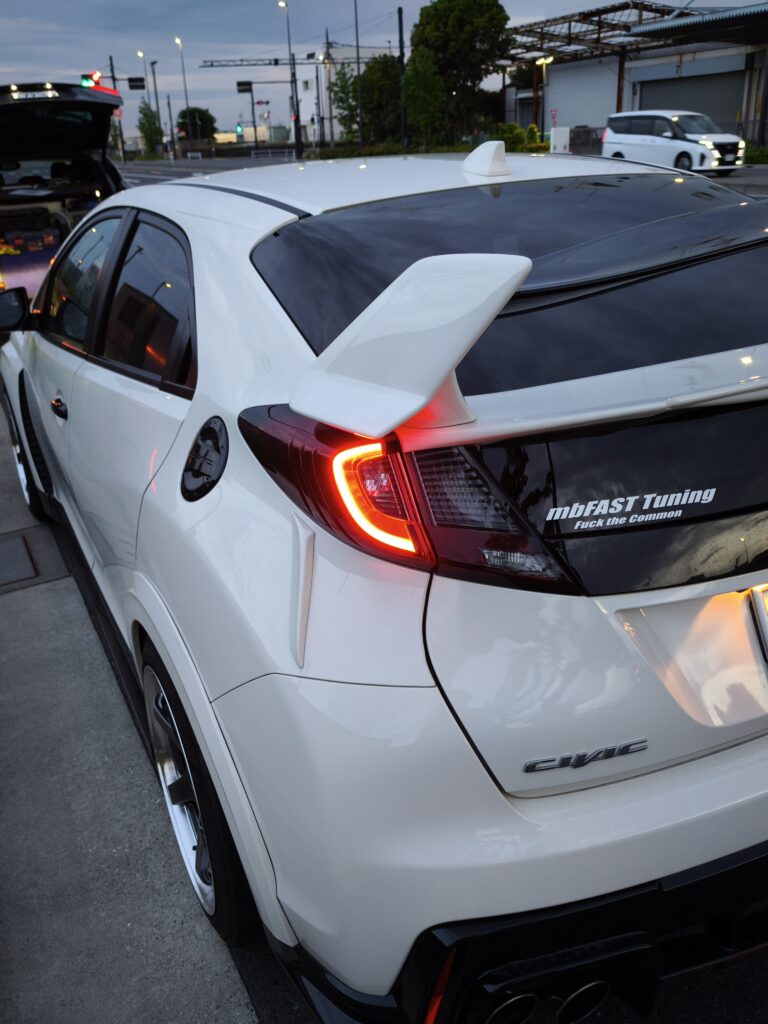 White hatchback viewed from the rear-left with a large aftermarket spoiler and red taillights, parked on a city street at dusk; decal reads mbFAST Tuning on the rear window/sill area.