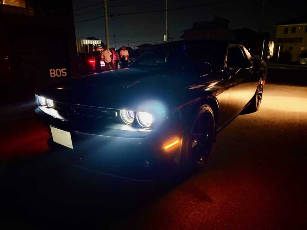 Nighttime street scene with a large black car in the foreground and a group of people gathered near another car in the background, illuminated by streetlights and car headlights.