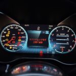Close-up of a car's illuminated digital instrument cluster with speedometer, tachometer, and warning icons in a dark cabin.