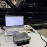 Laptop connected to automotive ECU tester on a workbench, with a black SUV in the background under a workshop ceiling.