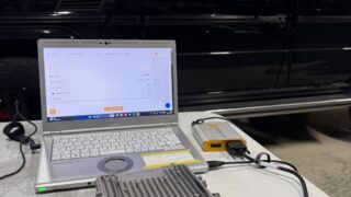 Laptop connected to automotive ECU tester on a workbench, with a black SUV in the background under a workshop ceiling.