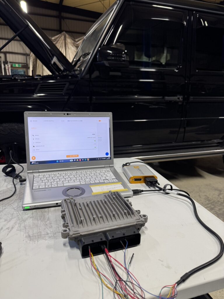 Laptop connected to automotive ECU tester on a workbench, with a black SUV in the background under a workshop ceiling.