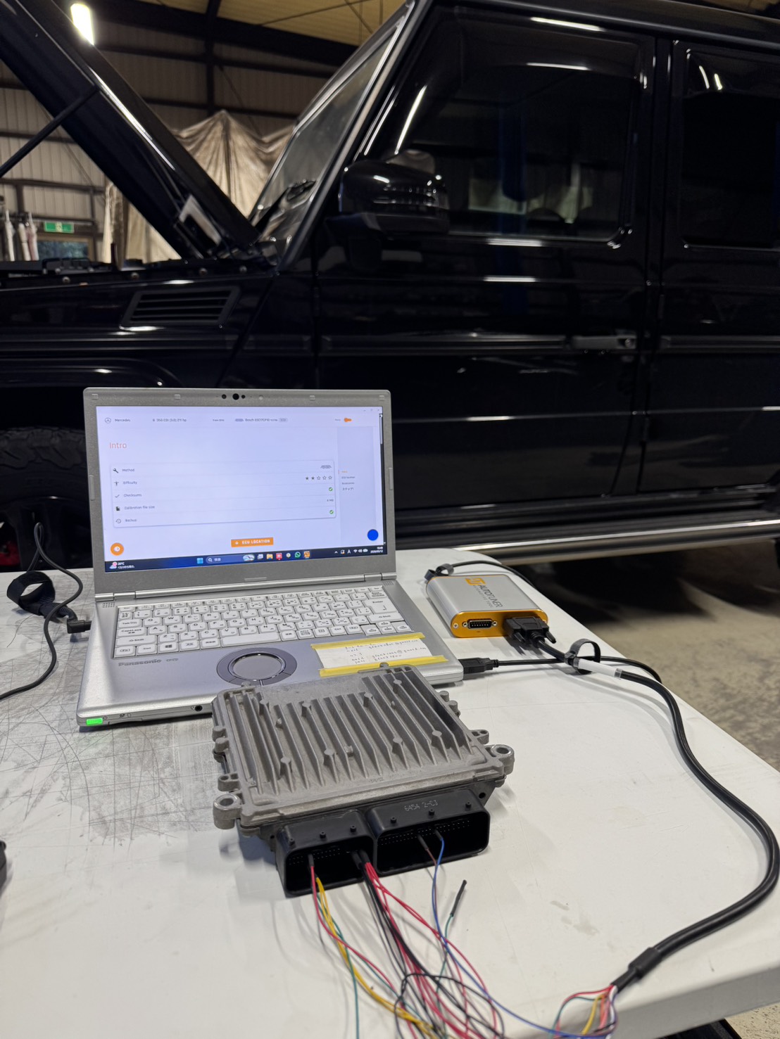 Laptop connected to automotive ECU tester on a workbench, with a black SUV in the background under a workshop ceiling.