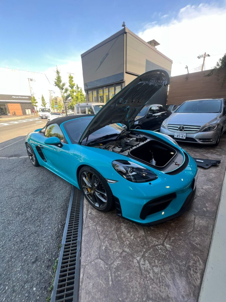 Blue sports car with its hood open in an outdoor parking area, showing engine bay and nearby vehicles and modern building in the background.
