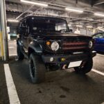 Black off-road SUV parked in a brightly lit indoor parking garage with a blue car nearby in the background.