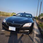 Front view of a black BMW parked on a road, showing the grille and headlight with a clear blue sky above.