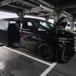 Black SUV with open hood parked in an industrial multi-level garage, front right wheel visible, reflective glass doors nearby, engine bay exposed under artificial lighting