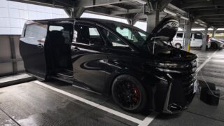 MVIMG_20260608_164910 – mbFAST Tuning Black SUV with open hood parked in an industrial multi-level garage, front right wheel visible, reflective glass doors nearby, engine bay exposed under artificial lighting