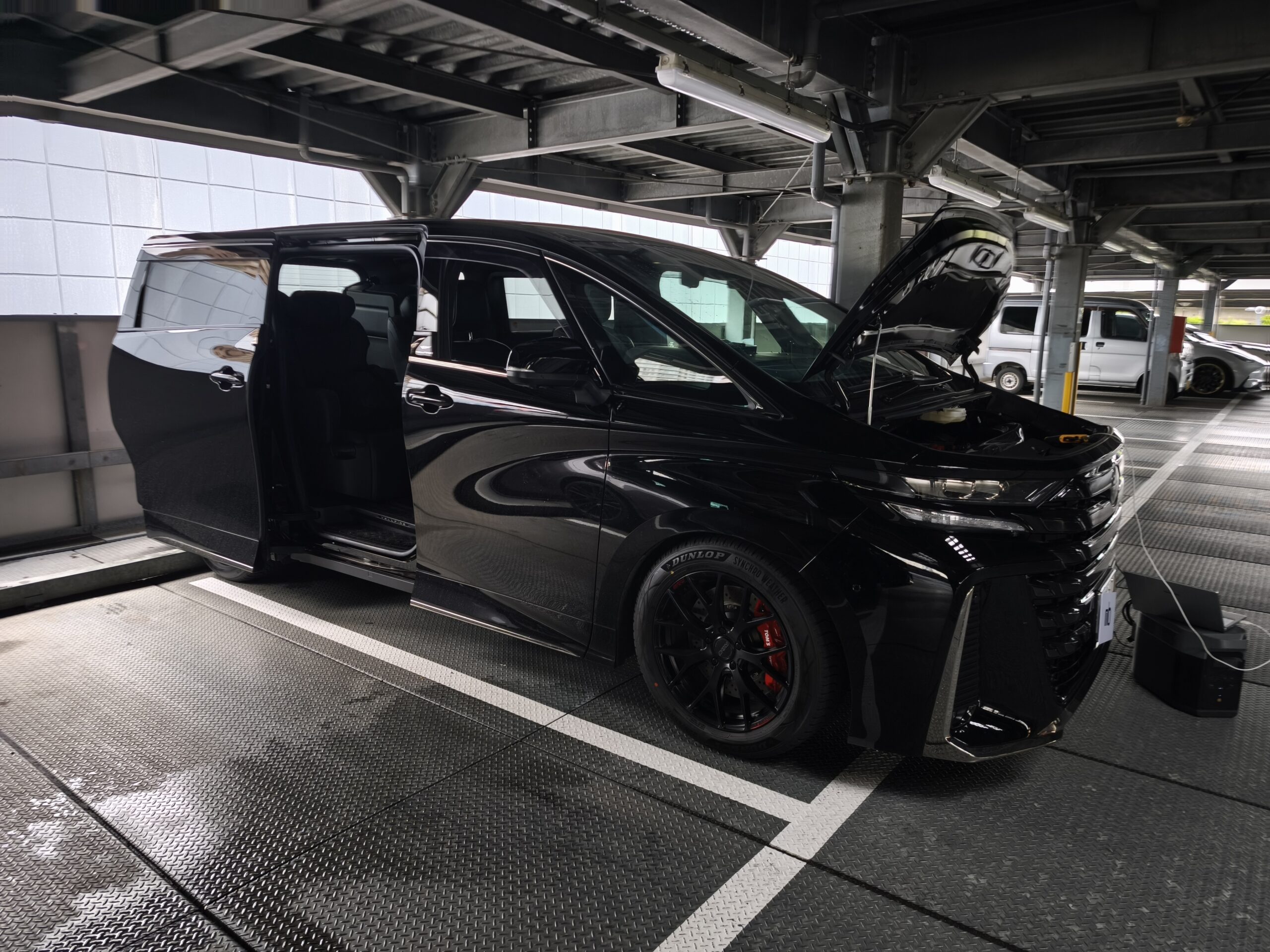 MVIMG_20260608_164910 – mbFAST Tuning Black SUV with open hood parked in an industrial multi-level garage, front right wheel visible, reflective glass doors nearby, engine bay exposed under artificial lighting