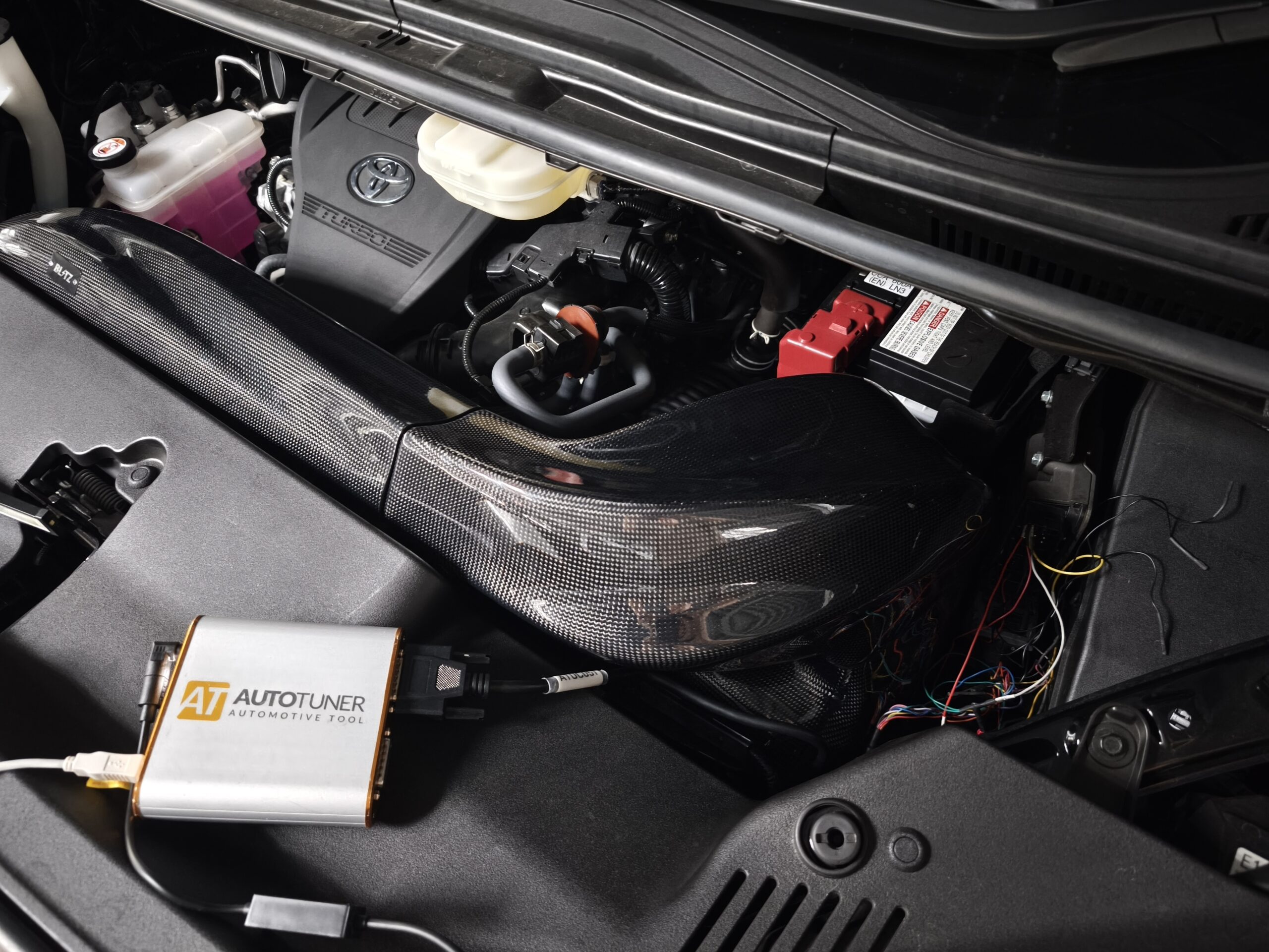 MVIMG_20260608_165117 – mbFAST Tuning Open engine bay showing coolant reservoir with pink liquid, engine cover with Toyota logo, and a boxed AT Autotuner device.