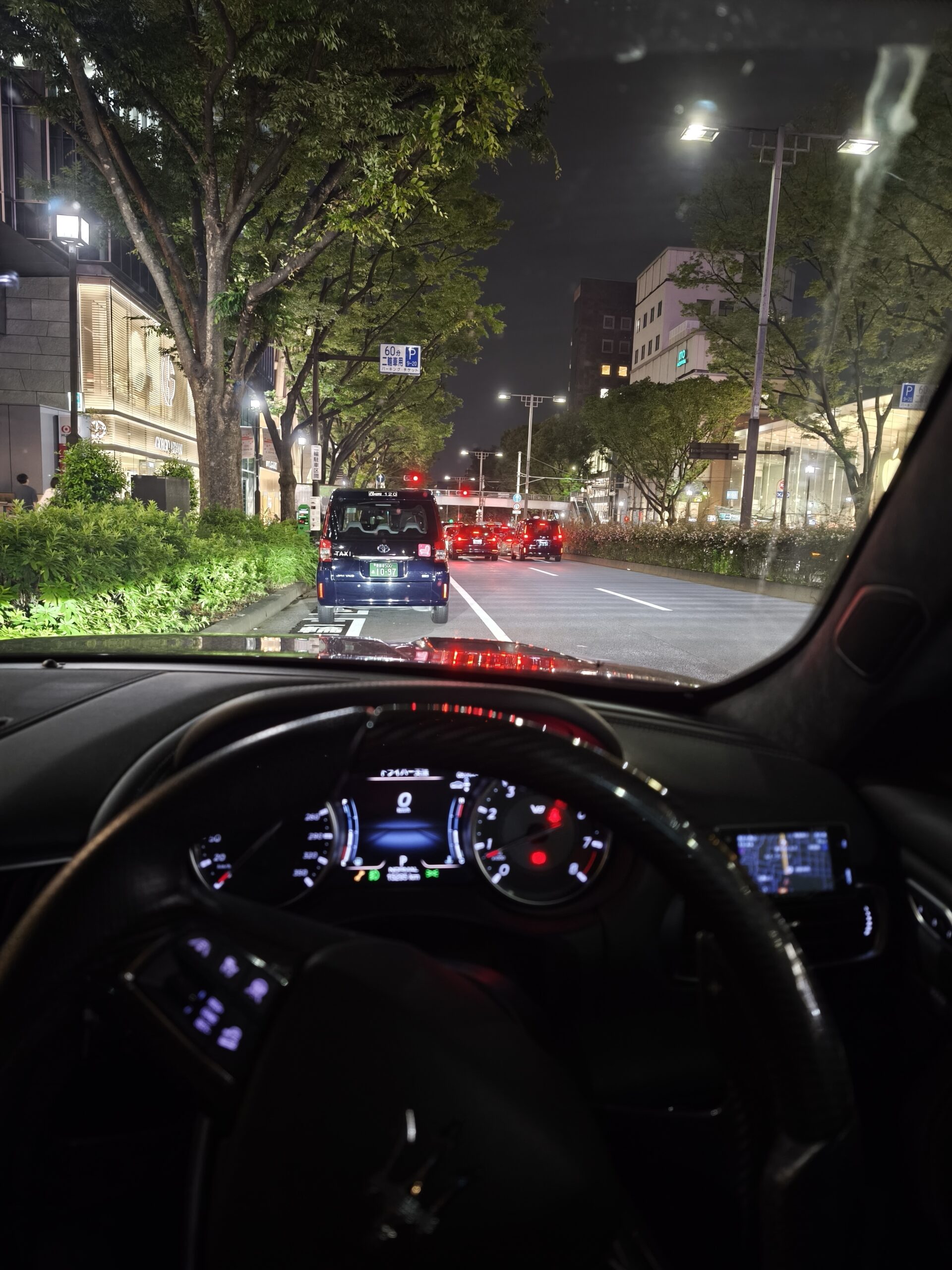 Nighttime city street view from inside a car, showing a line of vehicles ahead and illuminated storefronts along a tree-lined boulevard; dashboard in foreground.