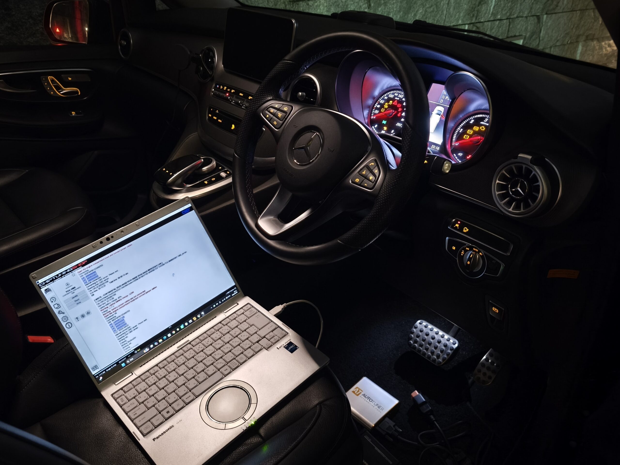 Inside a luxury car cockpit at night: steering wheel, illuminated dashboard, center console, and a laptop on the passenger seat