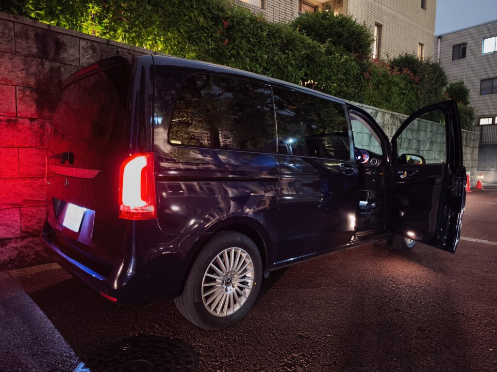 Black van parked along a brick wall at night with its side door open, red taillights glowing and wet asphalt visible.