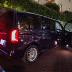 Black van parked along a brick wall at night with its side door open, red taillights glowing and wet asphalt visible.