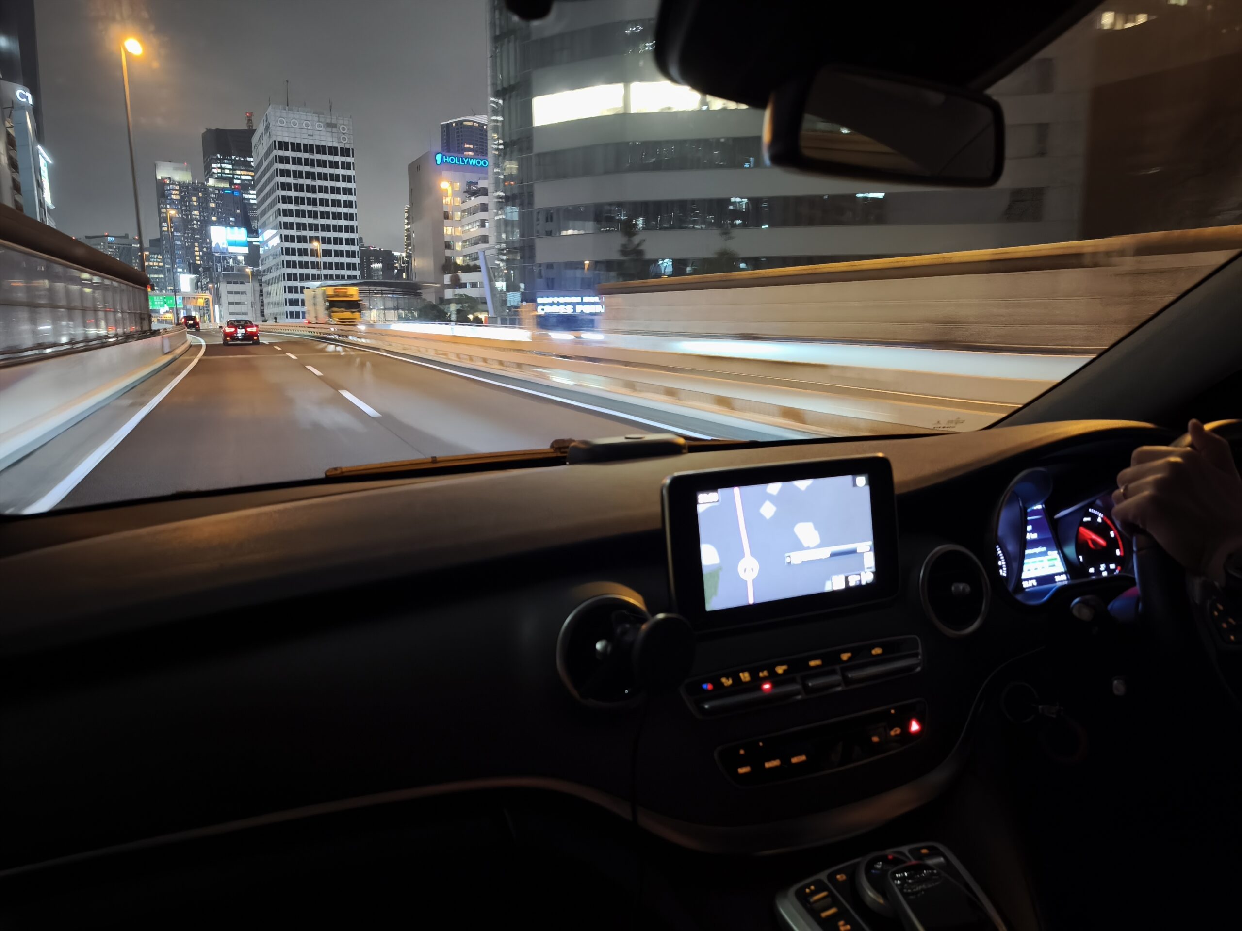 Nighttime city highway viewed from the car’s driver seat, with the dashboard and navigation screen in the foreground.