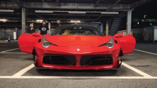 Front view of a bright red sports car with its doors open in an underground parking garage, with three people standing behind the car.
