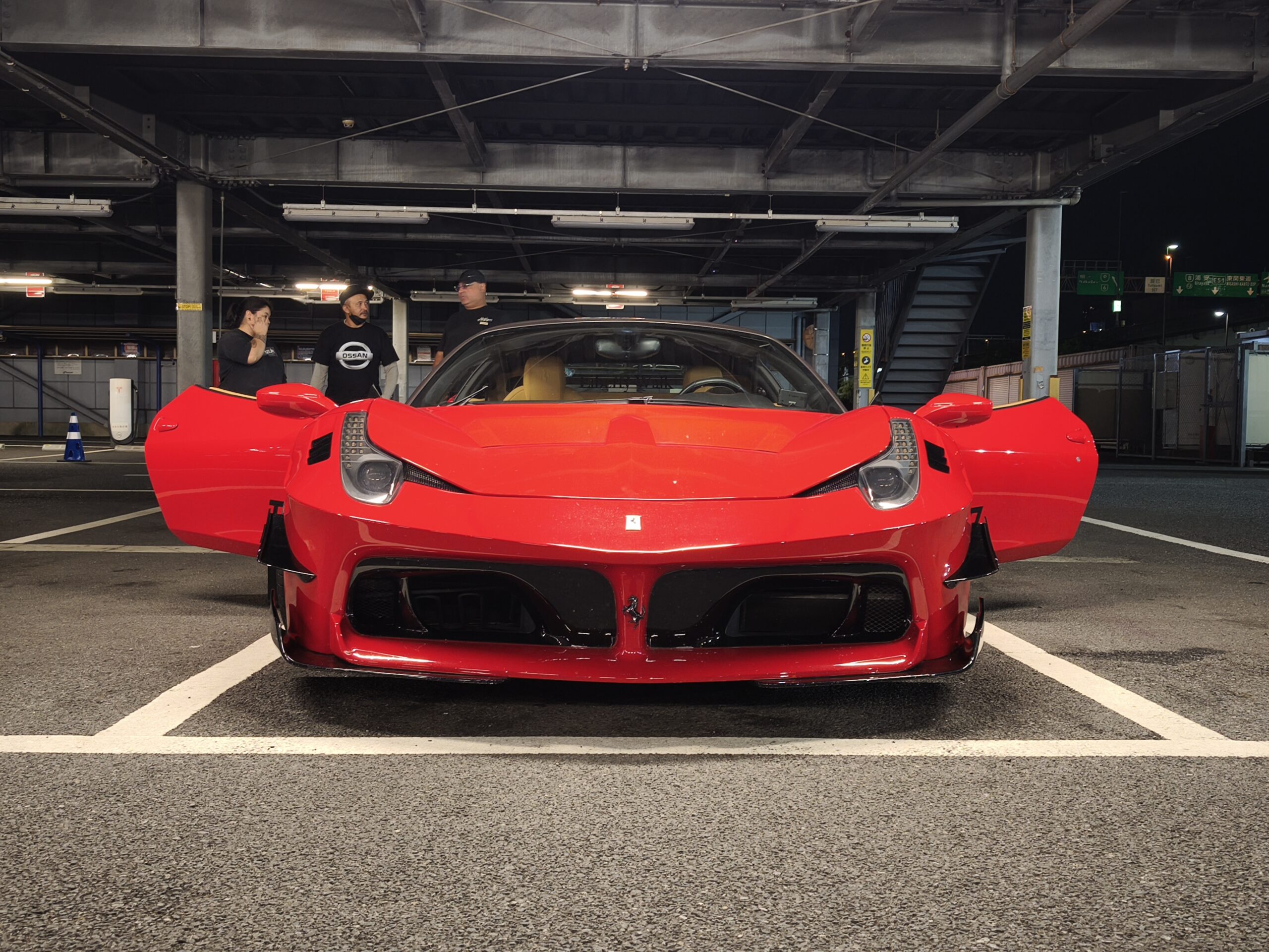 Front view of a bright red sports car with its doors open in an underground parking garage, with three people standing behind the car.