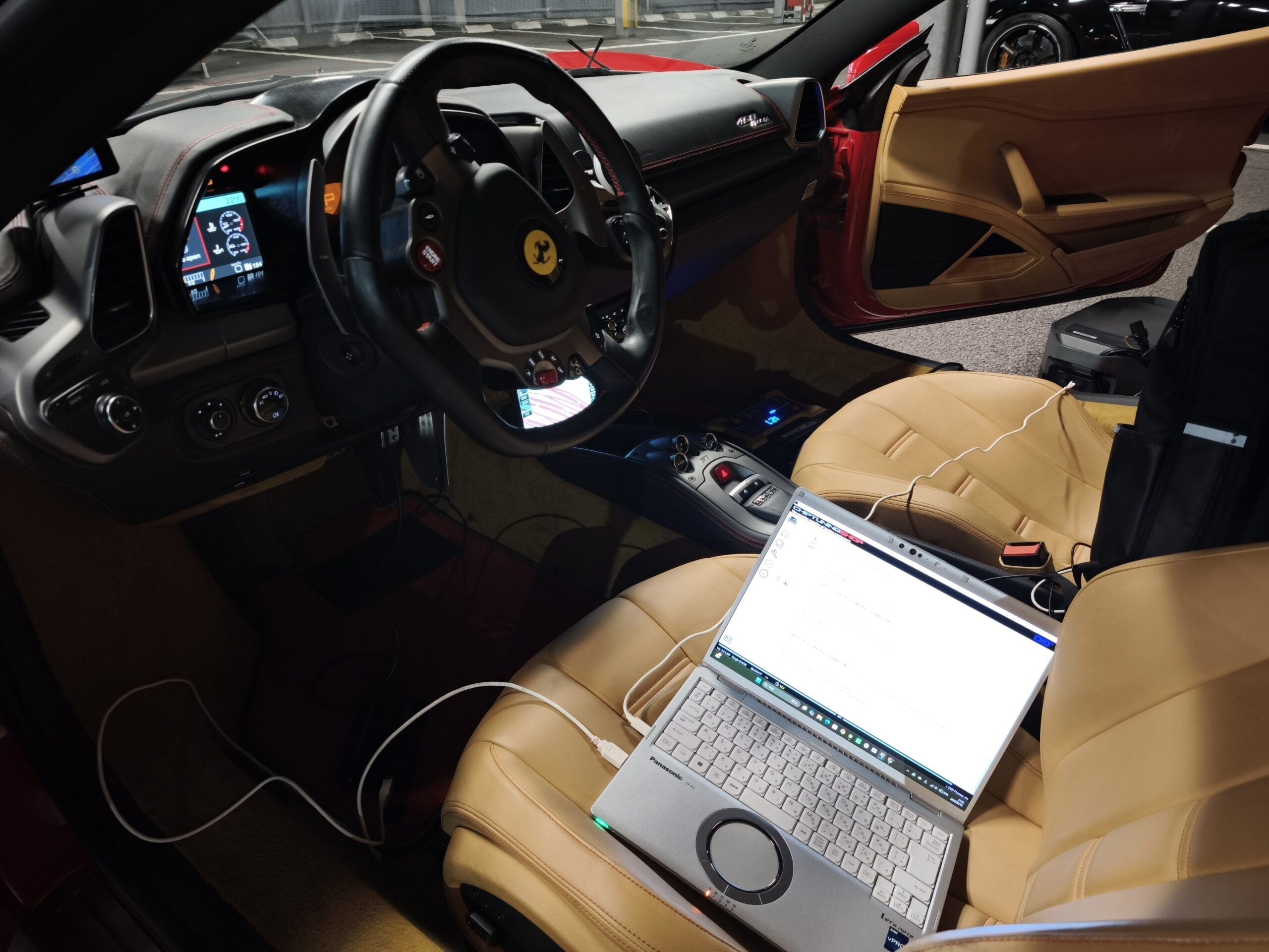 Interior of a high-end car with tan leather seats, steering wheel with logo, and a laptop on the passenger seat connected by charging cables in the foreground.