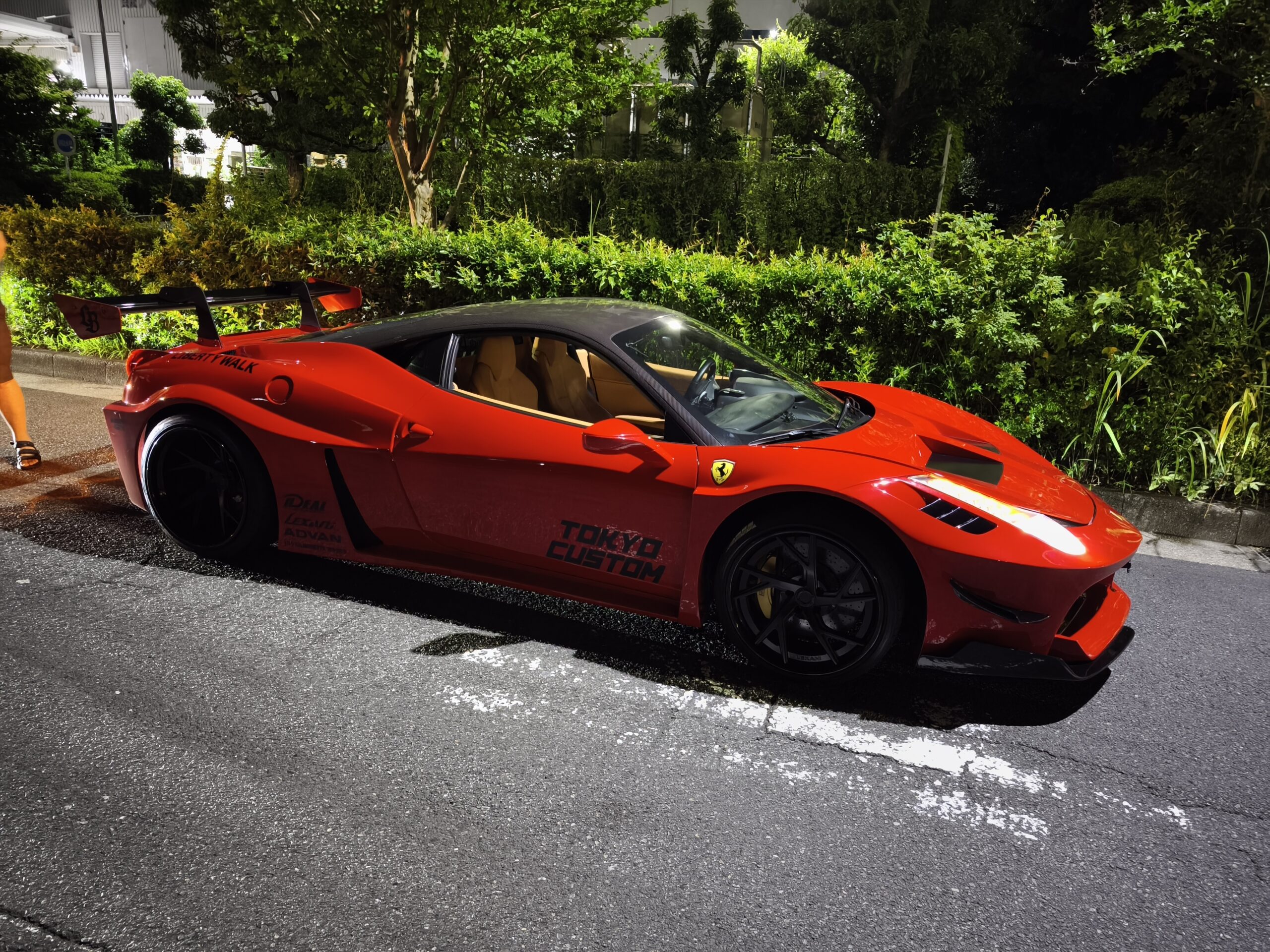 Red Ferrari sports car parked on a street at night with green hedges and trees in the background and a glossy red exterior.